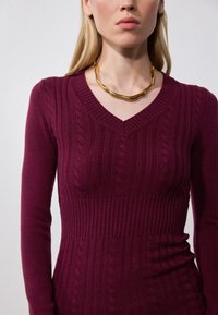 Pull en tricot bourgogne avec un décolleté en V, présentant des côtes texturées et des motifs en câble ; associé à un collier doré en forme de vague.