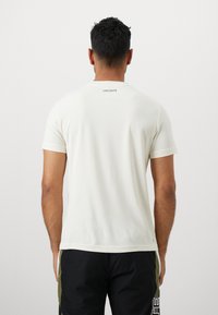 Camiseta blanca de manga corta con una textura suave, que presenta un pequeño logo negro de "LACOSTE" en la parte superior de la espalda. Diseño clásico de crew neck.