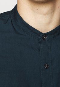 Ciemnozielona koszula z kołnierzykiem mandaryńskim, zapięciem na guziki oraz teksturowanym materiałem z subtelnymi liniami. Guzik jest okrągły i czarny.