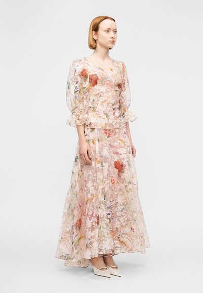 Robe longue à imprimé floral dans des tons pastel doux, avec tissu transparent, manches ballon et jupe fluide. Associée à des chaussures blanches à talons.