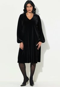 Robe en velours noir avec un décolleté en V et des manches longues. Présente un détail floral à l'épaule et arrive au-dessus du genou. Portée avec des talons noirs.