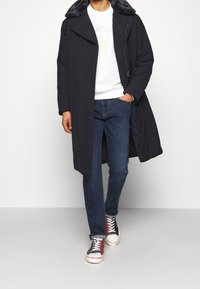 Person med en mörk lång kappa med pälskrage, vit sweatshirt med björngrafik, blå jeans och flerfärgade högklackade sneakers.