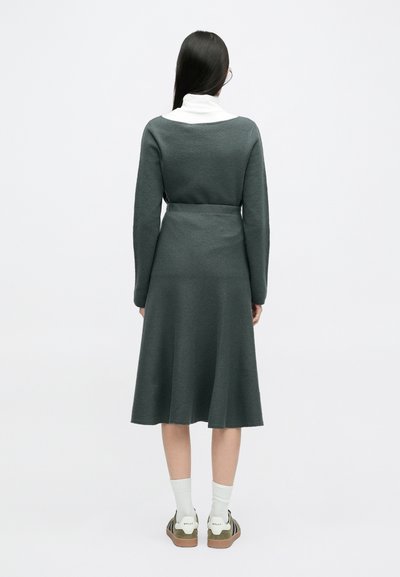 Μακρυμάνικο, σκούρο πράσινο φόρεμα με στενή μέση, φουσκωτή φούστα και μαλακή υφή. Φορεμένο πάνω από ένα λευκό πουλόβερ, συνδυασμένο με αθλητικά παπούτσια και κάλτσες.