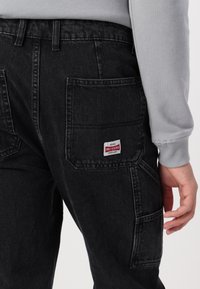 Sorte denim bukser med en afslappet pasform, der har store sidelommer og en rød og hvid mærketikette bagpå. Blød tekstur, synlige syninger.