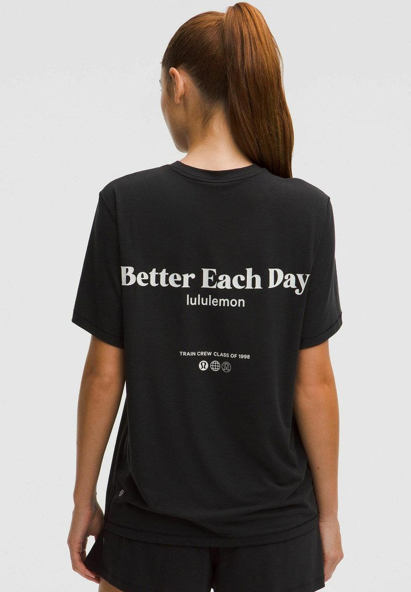 Hosszú vörös lófarokkal rendelkező nő, fekete laza pólót visel, amelyen az áll: "Better Each Day lululemon", és fekete rövidnadrágot, háttal áll.