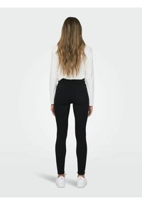 Femme aux longs cheveux ondulés, portant un haut blanc à manches longues, un pantalon noir ajusté et des baskets blanches, debout de dos sur un fond uni.