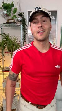Červené tričko Adidas s bílými pruhy na rukávech, nošené s béžovými kalhotami. Doplňky zahrnují béžovou čepici a viditelné tetování na paži.