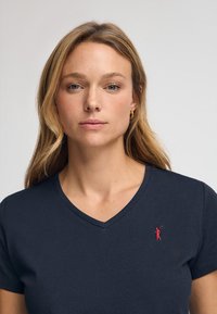 Marinblå kortärmad t-shirt med v-neck i bomull; har en liten röd broderad logotyp på vänster sida. Slät textur.