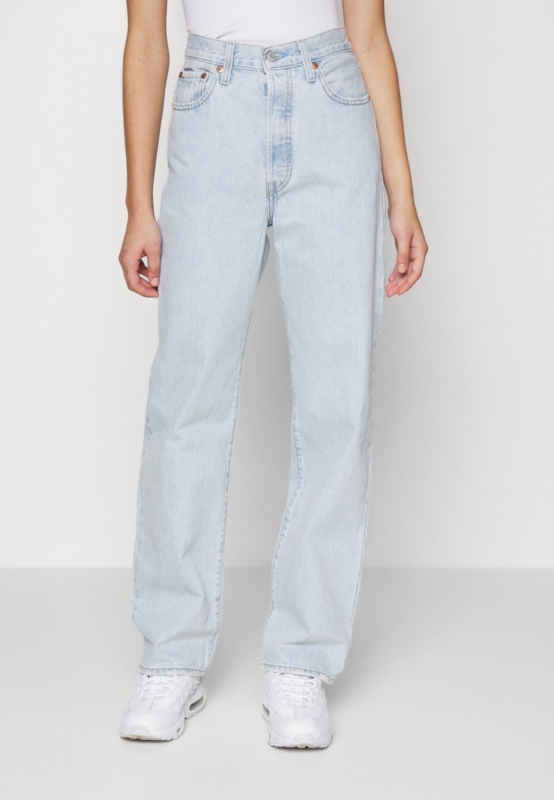 Personne portant un jean droit taille haute bleu clair et des baskets blanches, debout devant un fond clair uni.