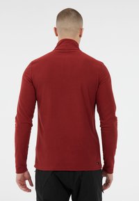 Camicia rossa a maniche lunghe con colletto alto, realizzata in tessuto morbido e elastico. Presenta un'orlo dritto e una comunicazione pubblicitaria minima sul lato.