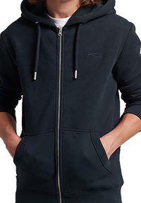 Felpa con cappuccio navy con zip in morbido misto cotone; presenta cappuccio con coulisse, tasche frontali e logo in rilievo sul petto.