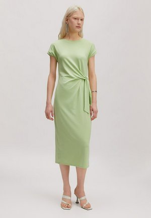 Femme aux longs cheveux blonds portant une robe midi vert clair avec un nœud sur le côté et des sandales blanches à talons, debout devant un fond uni.