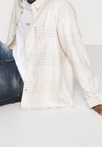 Crème geruite blouse met lichtbeige patronen, knoopsluiting aan de voorkant en één borstzak, gecombineerd met donkere versleten jeans.