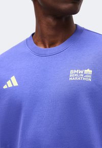 Gros plan d'une personne portant un t-shirt violet à col rond avec un logo Adidas jaune, le texte BMW Berlin Marathon et un graphisme de la porte de Brandebourg.