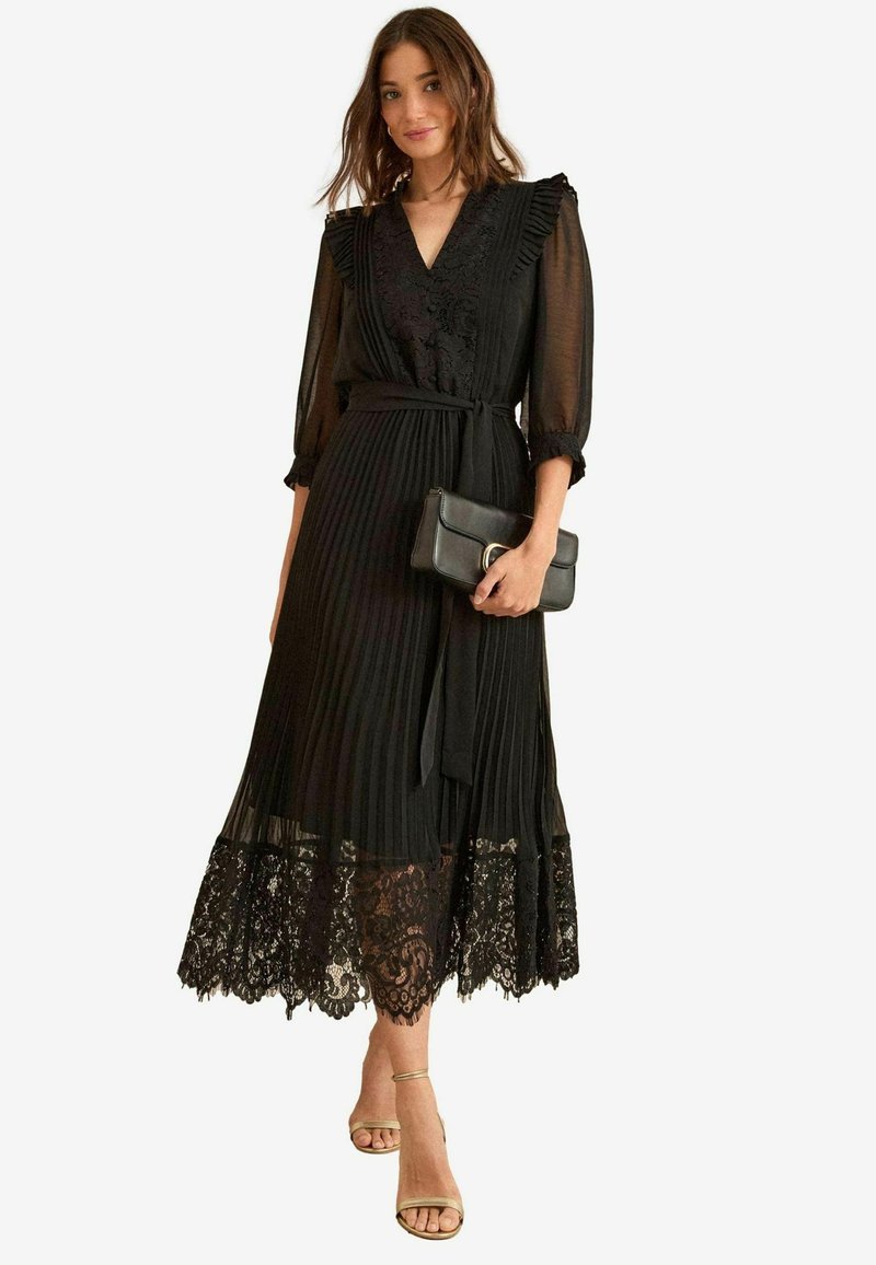 Robe plissée noire avec un décolleté en V profond, des détails en dentelle à l'ourlet et une ceinture à la taille. Manches longues transparentes avec des volants. Accentuée par une pochette.