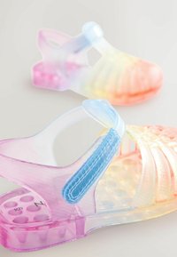 Permatomos želatininės basutės su gradientiniu spalvos perėjimu nuo rožinės iki mėlynos, tekstūruotos juostos ir raštuota padų dalimi su apskritais skylutėmis.