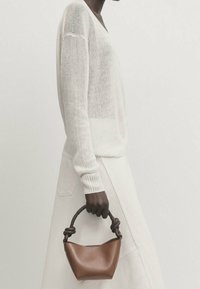 Brązowa skórzana torebka z węzłowym, teksturowanym uchwytem. Modelka w luźnym białym swetrze i kremowej spódnicy, widok z boku.