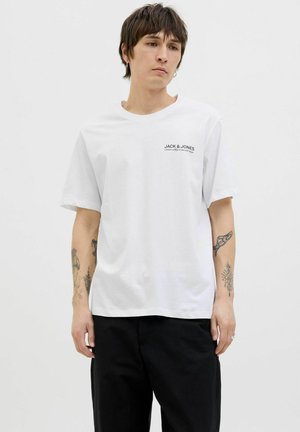 Hvid bomulds t-shirt med rund hals, løs pasform, med et lille sort logo på venstre bryst. Båret med sorte bukser.