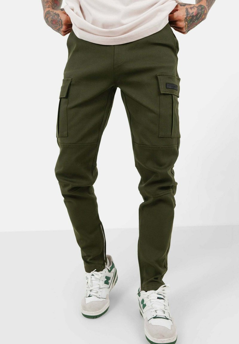 Olijf groene cargobroek met taps toelopende pasvorm, voorzien van twee zijzakken, een gladde stof en een patchlabel op de voorkant. Gepaard met witte sneakers.
