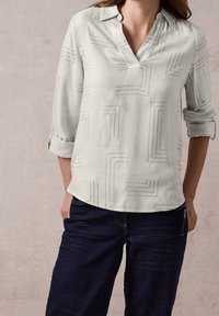Blouse gris clair à col en V, présentant des motifs géométriques en lignes noires. Manches longues retroussées, en tissu doux, associée à un jean foncé.