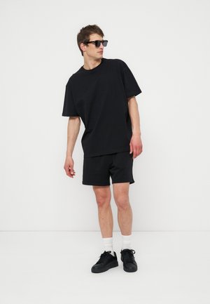 Homme portant un t-shirt noir oversize, un short noir, des baskets noires, des chaussettes blanches et des lunettes de soleil, debout devant un fond blanc uni.