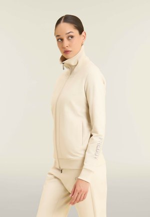 Giacca zip-up beige con colletto alto, tasche laterali e un'accaento testurizzato sulla manica. Tessuto liscio con un design aderente.