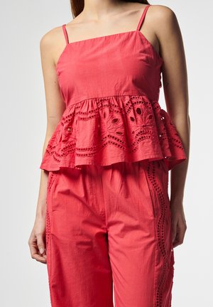 Frau mit rotem ärmellosem Oberteil mit Spitzen-Details und passenden roten Hosen mit seitlichen Spitzenakzenten vor einfarbigem Hintergrund.
