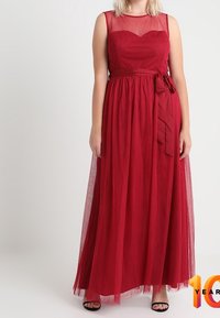 Röd, ärmlös maxiklänning med meshparti, figurnära överdel och flödande tärnkjol, inklusive ett knytband i midjan. Svarta skor synliga.