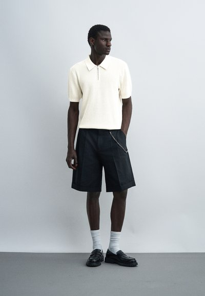 Homme portant un polo blanc à manches courtes zippé, un short noir jusqu'au genou avec une chaîne, des chaussettes blanches et des mocassins noirs, debout contre un mur uni.