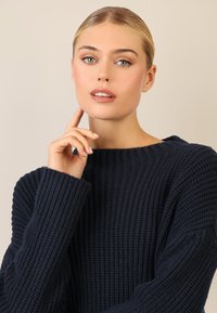 Granatowy sweter dzianinowy z ryżowym splotem i szerokim dekoltem, z długimi rękawami i luźnym krojem. Modelka trzyma dłoń na brodzie.