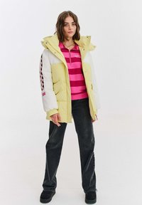 Žltá a biela pufková bunda s textúrovaným povrchom, vybavená kapucňou a ružovými akcentmi so vzorom, v páre s ružovým prúžkovaným tričkom a čiernymi nohavicami.