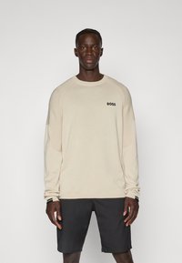 Hellbeige Pullover mit langen Ärmeln und gerippten Schulterdetails sowie einem schwarzen "BOSS"-Logo auf der Brust, kombiniert mit dunklen Shorts.