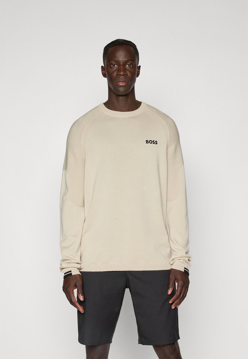 Hellbeige Pullover mit langen Ärmeln und gerippten Schulterdetails sowie einem schwarzen "BOSS"-Logo auf der Brust, kombiniert mit dunklen Shorts.
