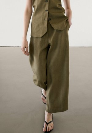 Personne portant un pantalon ample couleur vert olive et un haut sans manches assorti à boutons, associé à des sandales noires à lanières, debout sur un sol gris.