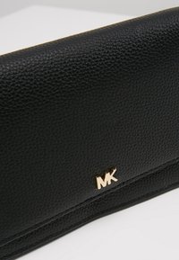 Cartera de cuero negro texturizado con un logo dorado de MK, un diseño elegante con un acabado suave y un sutil detalle de costura a lo largo de los bordes.