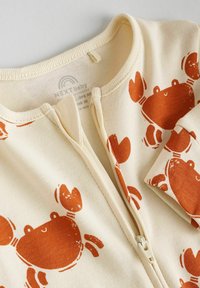 Cream-colored baby jumpsuit featuring a front zipper and orange crab motifs. Material is soft with a smooth texture, suitable for infants.