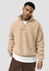 Beige fleece-genser med høy krage, glidelås og en subtil logo-detail. Myk tekstur og avslappet passform, lagd over en hvit skjorte.