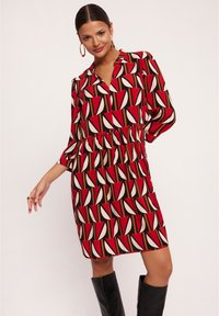 Czerwona i czarna sukienka z geometrycznym wzorem, z kołnierzykiem, długimi rękawami oraz wcięciem w talii. Teksturowana tkanina z wyraźnymi kształtami.