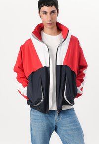 Lett vindjakke i rød, hvit og marineblå. Har høy krage, glidelås og sidelommer. Glatt tekstur og sporty design.