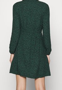 Robe à imprimé floral vert foncé avec des manches longues, un col et une taille ajustée. Présente une jupe évasée et des détails floraux noirs partout.