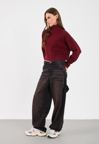 Burgunderfarbener Rollkragenpullover mit geripptem Saum, kombiniert mit lockeren schwarzen Jeans und klobigen, mehrfarbigen Turnschuhen.