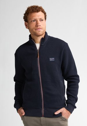 Veste zippée bleu foncé avec un motif en maille texturé, col haut et poignets côtelés. Présente un écusson de logo et des poches latérales.