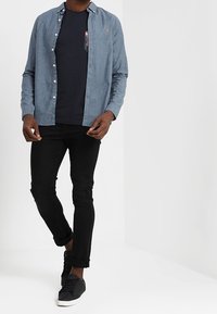 Světle modrá košile na knoflíky přes tmavé tričko s květinovou kapsou; doplněno černými slim-fit džíny a černými teniskami.