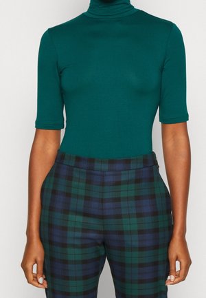 Vrouw die een groene coltrui met halve mouwen draagt en een blauwgroene geruite hoge taille broek, staand tegen een effen achtergrond.