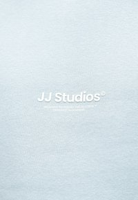 Gaiši zils kokvilnas džemperis ar uzrakstu "JJ Studios" biezos baltos burtiem, blakus ar mazākiem burtiem citas zīmola detaļas.