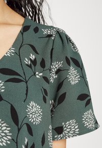 Zelená texturovaná látka s květinovým vzorem v bílé a černé. Krátké nabírané rukávy a design s V-výstřihem.