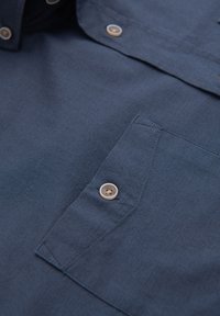 Tissu de chemise bleu foncé avec un gros plan sur une poche poitrine avec un bouton et des coutures détaillées.