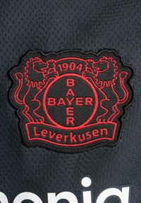 Parche de tela negra con el logotipo del Bayer Leverkusen, con leones rojos bordados y texto "1904" y "BAYER Leverkusen." Fondo con textura.