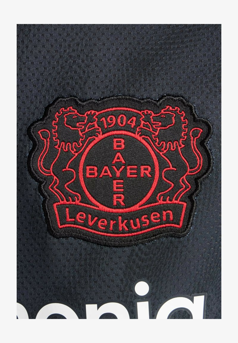 Schwarzer Stoffaufnäher mit dem Logo von Bayer Leverkusen, mit roten, gestickten Löwen und den Schriftzügen „1904“ und „BAYER Leverkusen“. Strukturierter Hintergrund.