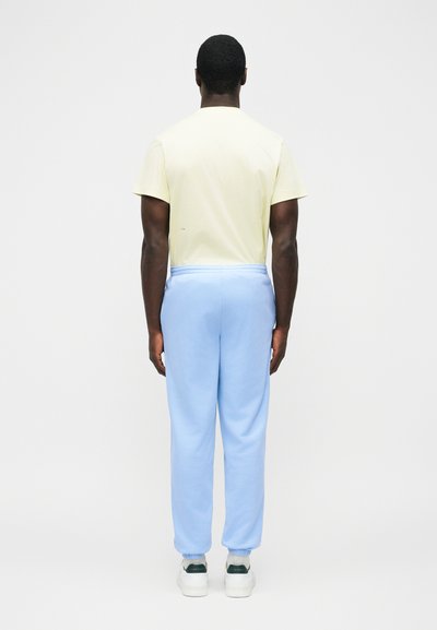 Egy férfi áll háttal, halványsárga rövid ujjú inget, világoskék tréningnadrágot, fehér zoknit és fehér sportcipőt visel, egyszínű háttér előtt.